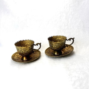May include: Two antique brass teacups and saucers. The cups and saucers have an ornate floral design and scalloped edges. The teacups have small handles. The saucers have a small pedestal base. The brass has a warm, golden hue.