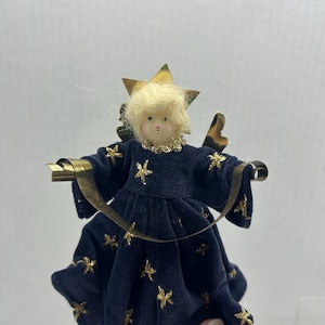 May include: A Christmas tree topper in the shape of an angel. The angel has a blonde head, a navy blue velvet dress with gold star accents, and gold wings. A gold star is on the angel's head.