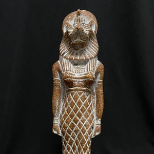 Könnte beinhalten: Eine braune und weiße Keramikstatue der ägyptischen Göttin Bastet, dargestellt als Frau mit einem Löwenkopf. Die Statue steht auf einem rechteckigen Sockel mit Hieroglyphen.