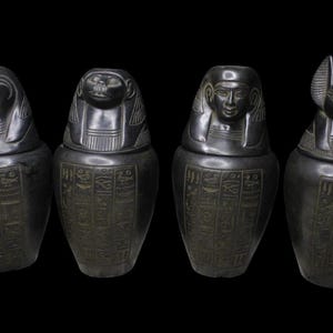 May include: A set of four black Egyptian canopic jars featuring carved lids representing a falcon, a baboon, a human, and a jackal. Each vessel is decorated with vertical columns of engraved hieroglyphic symbols across the main body surface.
