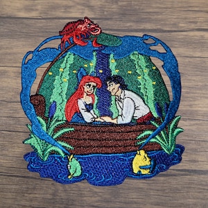 May include: Embroidered patch featuring a scene with a red-haired woman and a man in a boat, surrounded by blue water and green foliage. A red crab is perched above. The patch has a blue border and is set against a wooden background.