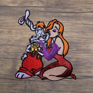 May include: Embroidered patch featuring a cartoon rabbit in a red outfit being kissed by a woman in a red dress and purple gloves. The rabbit is white and gray, and the woman has long orange hair. The patch is on a wooden surface.