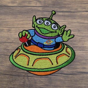 May include: Embroidered patch featuring a green alien character with three eyes, wearing a blue outfit and seated in a yellow and orange spaceship. The alien is smiling and waving. The patch is designed with detailed stitching.