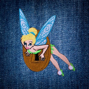 May include: An embroidered patch featuring a blonde fairy with blue wings emerging from a brown keyhole. The fairy has a green dress, light skin, and blue eyes. The patch is displayed against a dark blue denim fabric background with visible texture.