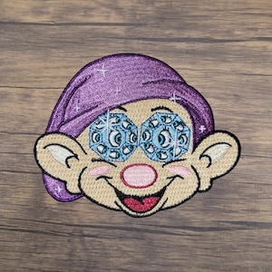 May include: Embroidered patch featuring a cartoon face with multiple eyes and a purple hat. The face has a tan complexion, a pink nose, and a smiling red mouth. The eyes are a light blue color with sparkling details. The patch is likely for clothing or accessories.