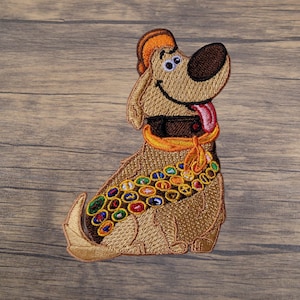 May include: Embroidered patch featuring a cartoon dog with a brown nose, orange hat, and a colorful collar. The dog is smiling with its tongue sticking out. The patch is designed with various colors and details.