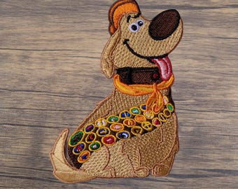 May include: Embroidered patch of a cartoon dog with a brown nose, orange hat, and a colourful collar. The dog is smiling with its tongue out. The patch is designed with various colours and details.