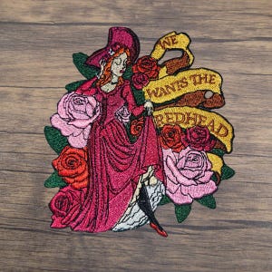 May include: Embroidered iron-on patch featuring a woman in a red dress and hat surrounded by roses. The banner reads "We Wants The Redhead".