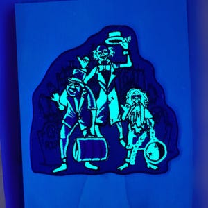 May include: A black and blue patch featuring three cartoon ghosts. The ghosts are wearing formal attire and are glowing a bright teal color. The background is a dark blue color.