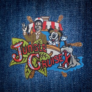 May include: Embroidered patch featuring the words "Jungle Cruise" in red, blue, and green, with images of Mickey Mouse, a boat, and a hippo. The patch is on a blue denim background.