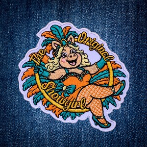 May include: An embroidered patch featuring a pig character dressed as a showgirl with orange and teal feathers, wearing a matching outfit and heels. The design is encircled by a gold ring with the text The Original Showgirl in cursive script.