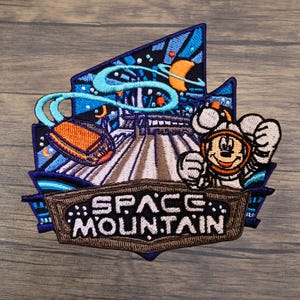May include: An embroidered patch featuring a space-themed scene with a rocket vehicle, a crescent moon, and a character wearing a space helmet. The text SPACE MOUNTAIN is stitched in white on a brown banner at the bottom of the dark blue design.