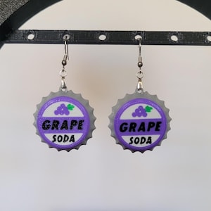May include: A pair of novelty earrings shaped like grape soda bottle caps. The earrings are silver-toned with a purple and white design. The text "GRAPE SODA" is printed on the front, along with a grape cluster graphic.