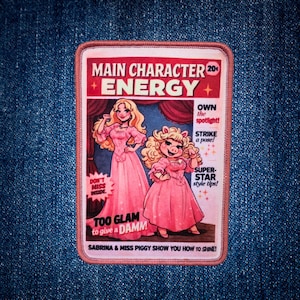 May include: A rectangular embroidered patch featuring a magazine cover design with the text MAIN CHARACTER ENERGY. It depicts a blonde woman and Miss Piggy in pink gowns. Additional text includes OWN THE SPOTLIGHT, STRIKE A POSE, and TOO GLAM TO GIVE A DAMN.