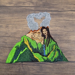 May include: Embroidered patch featuring two green volcanoes with a woman's face on one and a cartoon face on the other. The volcanoes are topped with gray smoke.