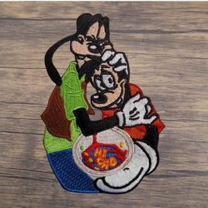 May include: Embroidered patch featuring the Disney characters Goofy and Max. Goofy is wearing a green shirt and brown vest, while Max is in a red shirt. The patch includes a bowl with colorful food. The patch is on a wooden surface.