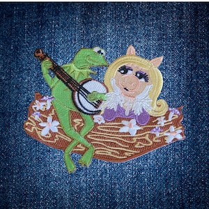 May include: Embroidered patch featuring Kermit the Frog playing a banjo for Miss Piggy. Kermit is green, and Miss Piggy is blonde with a purple dress. The patch is set on a brown base with white flowers, all on a blue denim background.