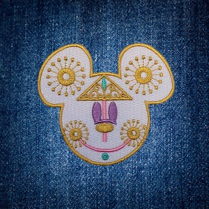May include: Embroidered patch featuring a stylized Mickey Mouse head with a Day of the Dead theme. The design includes white, gold, purple, and green elements, with floral accents and a decorative crown. The patch is set against a blue denim background.