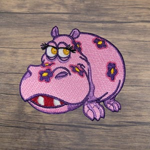 May include: Embroidered patch of a pink hippopotamus with purple eyelashes, a purple mouth, and purple flowers on its body.