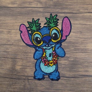 May include: Blue embroidered character with large eyes wearing yellow sunglasses and a colourful flower lei. The character has green pineapple on its head.