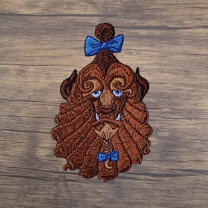 May include: Embroidered patch featuring a stylized brown Beast character with blue eyes and a blue bow. The patch has a detailed design with various shades of brown and blue, suitable for clothing or accessories.