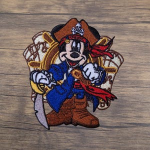 May include: Embroidered patch featuring a cartoon mouse dressed as a pirate. The mouse wears a brown pirate hat with a skull and crossbones, a red bandana, and a blue coat. He holds a sword and stands in front of a ship's wheel.