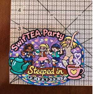 Puede incluir: Un parche bordado con cuatro figuras bailando, una tetera verde azulado y una taza rosa con vapor en forma de coraz&oacute;n. El texto dice "SWIFTEA Party" y "Steeped in FRIENDSHIP". Una regla muestra una anchura de 6,5 cm.