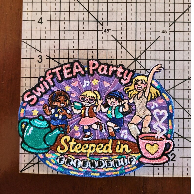 Puede incluir: Un parche bordado con cuatro figuras bailando, una tetera verde azulado y una taza rosa con vapor en forma de coraz&oacute;n. El texto dice "SWIFTEA Party" y "Steeped in FRIENDSHIP". Una regla muestra una anchura de 6,5 cm.
