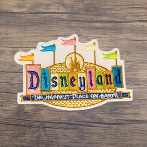 May include: Embroidered patch featuring the Disneyland logo with a castle and colorful flags. The text reads "Disneyland" in blue, pink, and yellow, with "THE HAPPIEST PLACE ON EARTH" below. The patch has a white border.