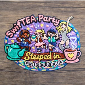 Puede incluir: Un parche bordado que muestra a cuatro figuras bailando sobre un fondo de rayos de sol morados. El texto dice "SwifTEA Party" y "Steeped in Friendship". Una tetera verde azulado est&aacute; a la izquierda y una taza rosa con un coraz&oacute;n a la derecha.