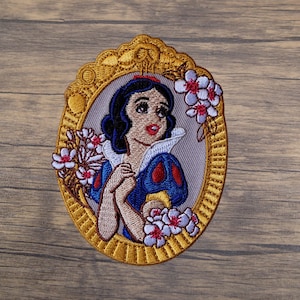 May include: Embroidered patch featuring Snow White in a gold oval frame with floral accents. The patch showcases Snow White with black hair, a red bow, and a blue and white dress. The frame and flowers have gold and white details.