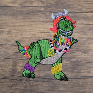 May include: Embroidered patch featuring a green dinosaur with a Viking helmet, orange hair, and a colorful lei. The dinosaur wears purple and yellow armbands and a peace sign. The word "REX" is on the lei.