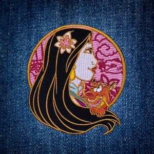 May include: A circular embroidered patch featuring a profile view of a person with long black hair, a pink flower accessory, and a small red dragon character. The design is set against a pink background with a gold border, stitched onto blue denim.