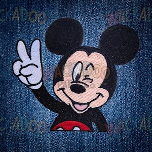 May include: Embroidered patch featuring Mickey Mouse giving a peace sign. The patch has a black outline, black ears, and a red bottom. Mickey's face is beige with black eyes and a black nose. The background is a blue denim.