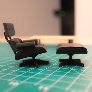 May include: Miniature black chair and ottoman set. The chair has a high back and armrests, and both pieces have a star-shaped base. The set is placed on a green grid surface.