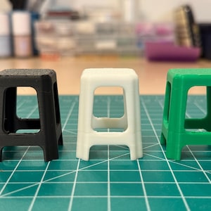 May include: Three miniature stools in black, white, and green are arranged in a row. Each stool has a square seat and four rectangular legs, with a rectangular opening in the center. The stools are on a green surface.