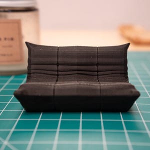 Op de afbeelding: Een kleine, zwarte, miniatuur bank met een geometrisch ontwerp. De bank is gemaakt van een solide materiaal en heeft een lage rug met drie secties. Het staat op een groene snijmat.