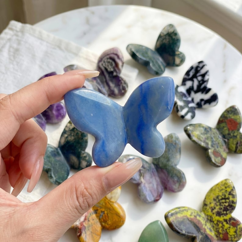 May include: A collection of gemstone butterfly figurines in various colors and patterns. The central butterfly is a light blue, while others display shades of green, purple, and yellow. The figurines are displayed on a white surface.