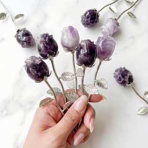 Peut inclure: Sculptures de roses en am&eacute;thyste. Les boutons de rose sont sculpt&eacute;s dans de la pierre violette et blanche, avec des tiges et des feuilles en m&eacute;tal argent&eacute;. Les roses sont dispos&eacute;es en bouquet, avec plusieurs autres &eacute;parpill&eacute;es.