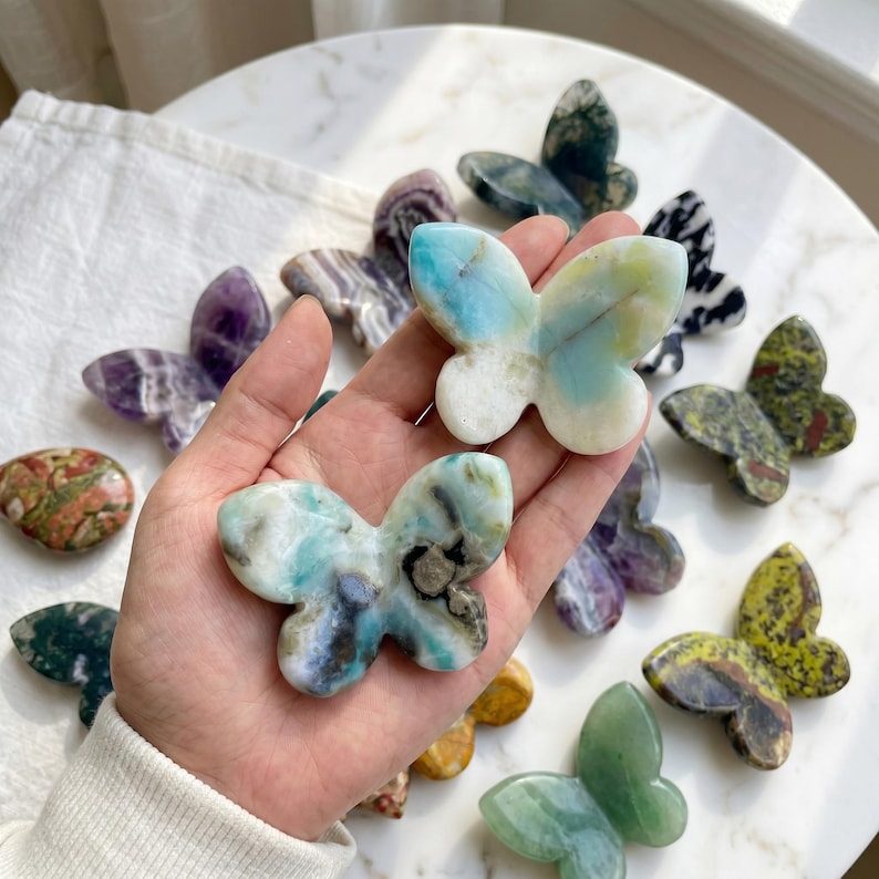 May include: A collection of butterfly-shaped stone carvings in various colors and patterns. The butterflies are held in a hand, with others scattered on a white surface. Colors include blue, green, purple, and brown.