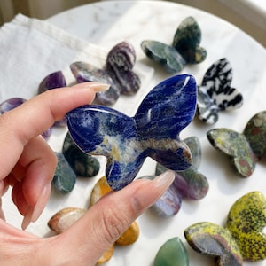 May include: A hand holds a blue butterfly-shaped crystal, showcasing its intricate patterns. Other butterfly crystals in various colors and patterns are scattered around, including green, purple, and yellow, creating a collection of decorative items.