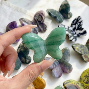 May include: A collection of carved stone butterfly figurines in various colors and patterns. A hand holds a green butterfly figurine with visible veining. Other butterflies are shades of purple, green, and black and white.