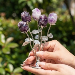 Peut inclure: Un bouquet d&eacute;coratif de roses en am&eacute;thyste, avec des boutons floraux violets et lavande. Les roses sont mont&eacute;es sur des tiges argent&eacute;es avec des d&eacute;tails de feuilles. L'arrangement est maintenu dans une base argent&eacute;e en forme de vase.