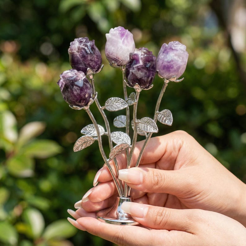 Peut inclure: Un bouquet d&eacute;coratif de roses en am&eacute;thyste, avec des boutons floraux violets et lavande. Les roses sont mont&eacute;es sur des tiges argent&eacute;es avec des d&eacute;tails de feuilles. L'arrangement est maintenu dans une base argent&eacute;e en forme de vase.