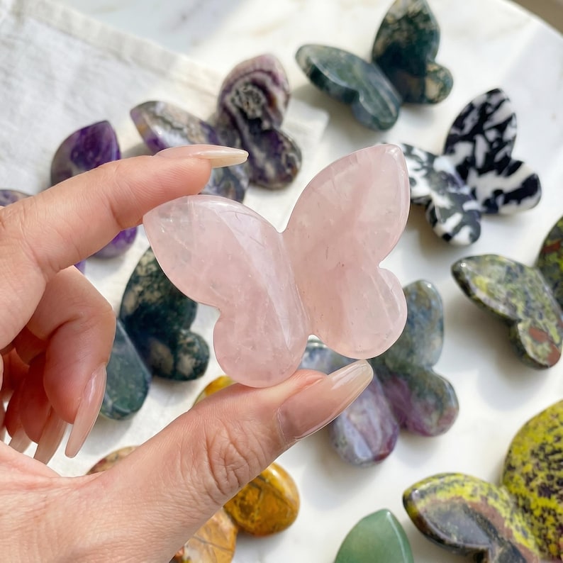 May include: A hand holds a pink rose quartz butterfly carving, surrounded by other butterfly-shaped stone carvings in various colors and patterns. The carvings are displayed on a white surface.