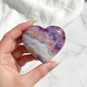 Puede incluir: Un cristal de amatista en forma de corazón con capas de tonos morados, blancos y anaranjados. La piedra pulida se sostiene en una mano, mostrando sus patrones y colores naturales. Una pieza decorativa para coleccionistas de cristales.