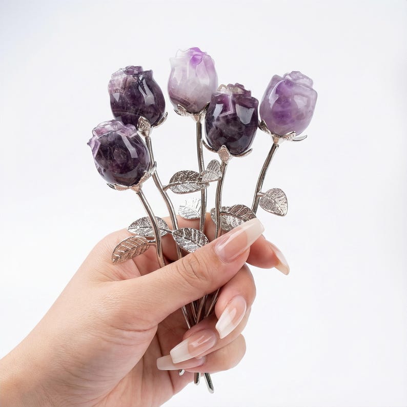 Peut inclure: Un bouquet de cinq sculptures de roses en am&eacute;thyste. Les boutons de rose sont sculpt&eacute;s dans une pierre pr&eacute;cieuse violette et blanche, avec des tiges et des feuilles argent&eacute;es. Les fleurs sont tenues dans une main sur fond blanc.