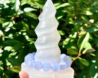 Puede incluir: Una escultura de cristal transparente en espiral con una pulsera de cuentas azul claro que descansa sobre su base. La escultura se sostiene frente a un fondo verde y frondoso, creando un contraste de texturas y colores. Las cuentas de la pulsera miden aproximadamente 1,3 cm de diámetro.