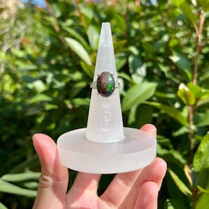 May include: A silver ring with a large, oval-shaped black opal gemstone. The opal displays vibrant green and blue flashes. The ring is displayed on a white, cone-shaped ring holder, with a green foliage background.