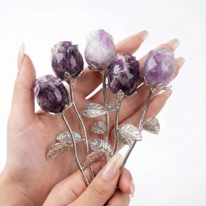 Peut inclure: Un bouquet de cinq sculptures de roses en am&eacute;thyste. Les boutons de rose sont un m&eacute;lange de violet fonc&eacute; et de blanc, avec des tiges et des feuilles argent&eacute;es. Les sculptures sont tenues dans une main sur fond blanc.